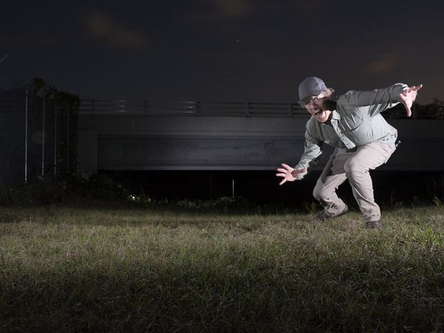De noche, un hombre se arrodilla y levanta los brazos a la cámara, frente al puente