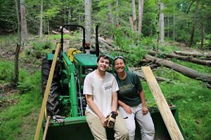 Two people sitting on a green tractor in a forested area smile at the camera. One is wearing a gray shirt and gloves, the other a green shirt with the year visible.