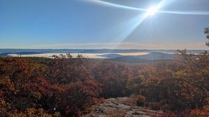 A scenic view of a sunlit landscape with rolling hills and autumn foliage. The sky is clear with a bright sun casting rays over the hills. A layer of fog or low clouds is visible in the valley.