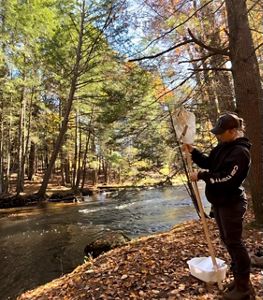 A person in a dark sweatshirt conducts research with a pole and attached equipment beside a stream in a forest with fall foliage.