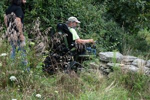 A person in an electric wheelchair navigates through a grassy area near a stack of stone slabs, assisted by another individual. The surroundings are lush with various plants and trees.
