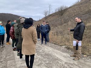 A man speaks to a group of people gathered around him at a restoration site. They are standing on a wide dirt track. A scrubby hill rises behind him.