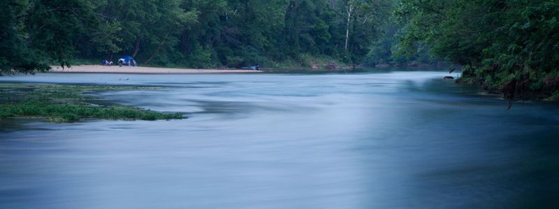 A wide river flows through a forest in the Ozarks of Missouri; a tent is set up on a sandy area along its banks.