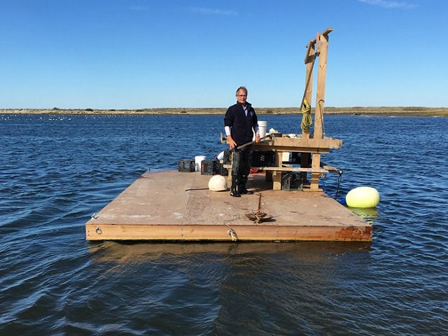 Oyster farmer Seth Garfield stands on a wooden, floating workspace in the middle of water.