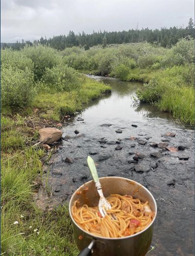 A pot of cooked spaghetti is held up in front of a winding steam that flows between grassy banks.