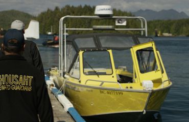 a man in a jacket that says, "ahousant stewardship guardians" walks away from the camera toward a boat