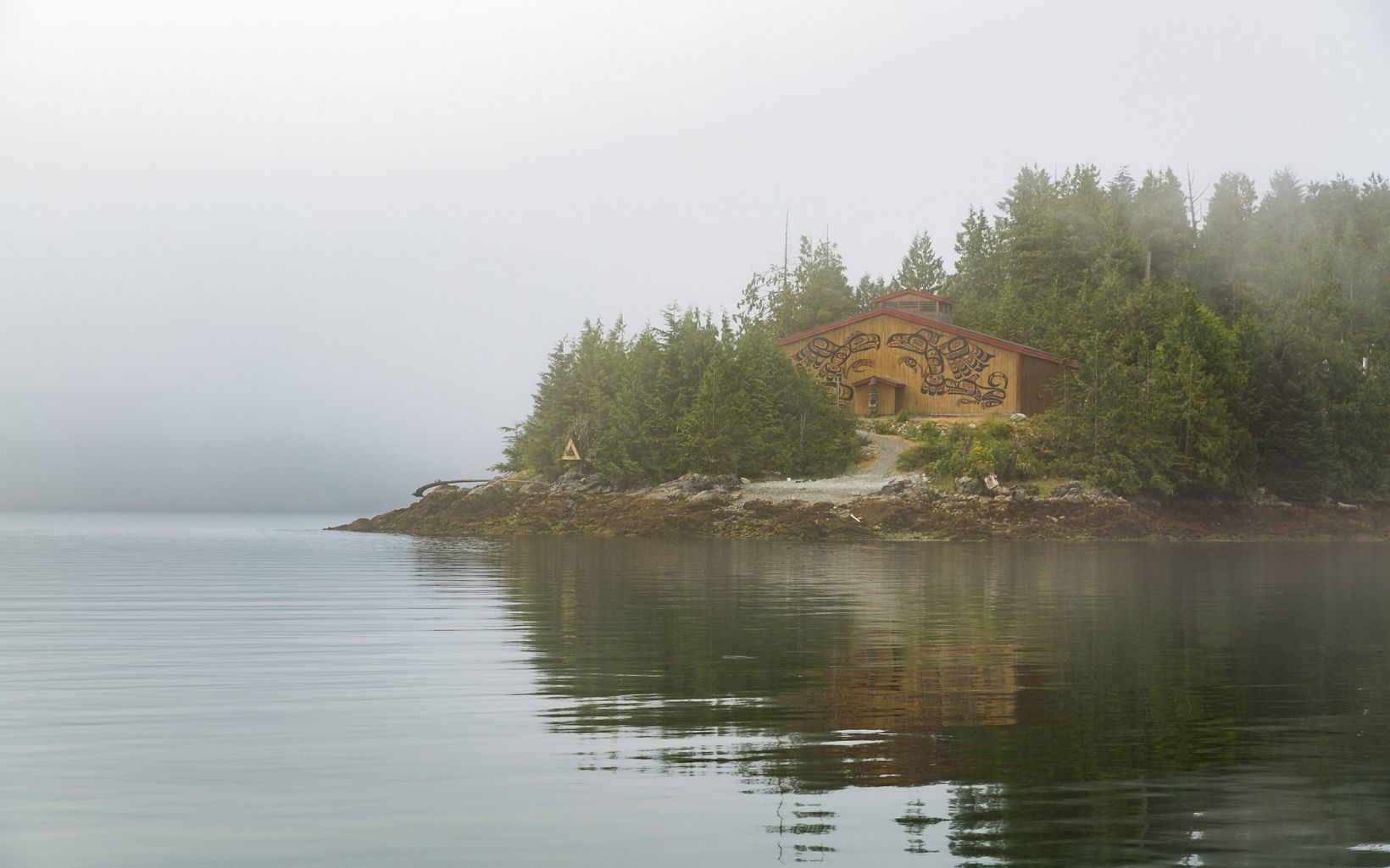 View of a small wooden building with Indigenous drawings on the front at the waterfront edge of a piece of land on a foggy day.