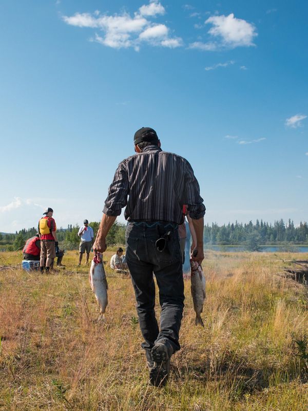 a man holding two fish walks away from the camera toward a group of people outside under a blue sky