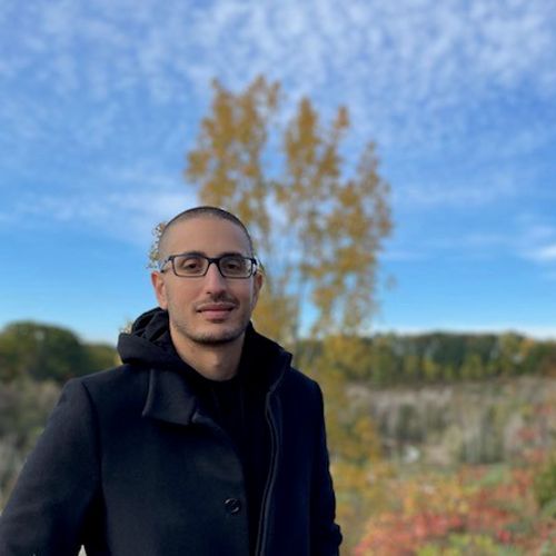 a man with glasses in a black jacket standing in front of trees