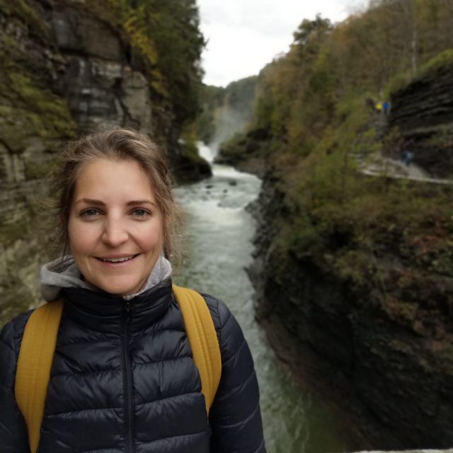 a smiling woman in a down jacket standing in front of a waterfall