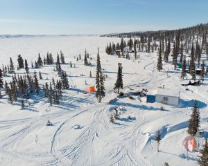 aerial view of a snow-covered landscape with snowmobile tracks, a cabin, a few people, and trees