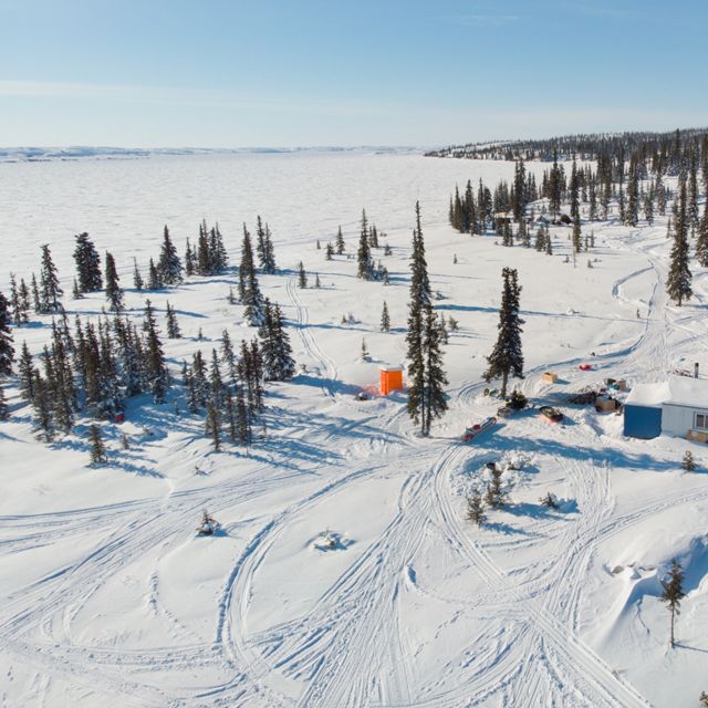aerial view of a snow-covered landscape with snowmobile tracks, a cabin, a few people, and trees