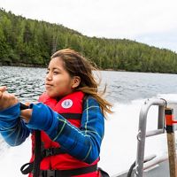 a young girl in a red life jacket holds onto a line on a boat going past a green shoreline