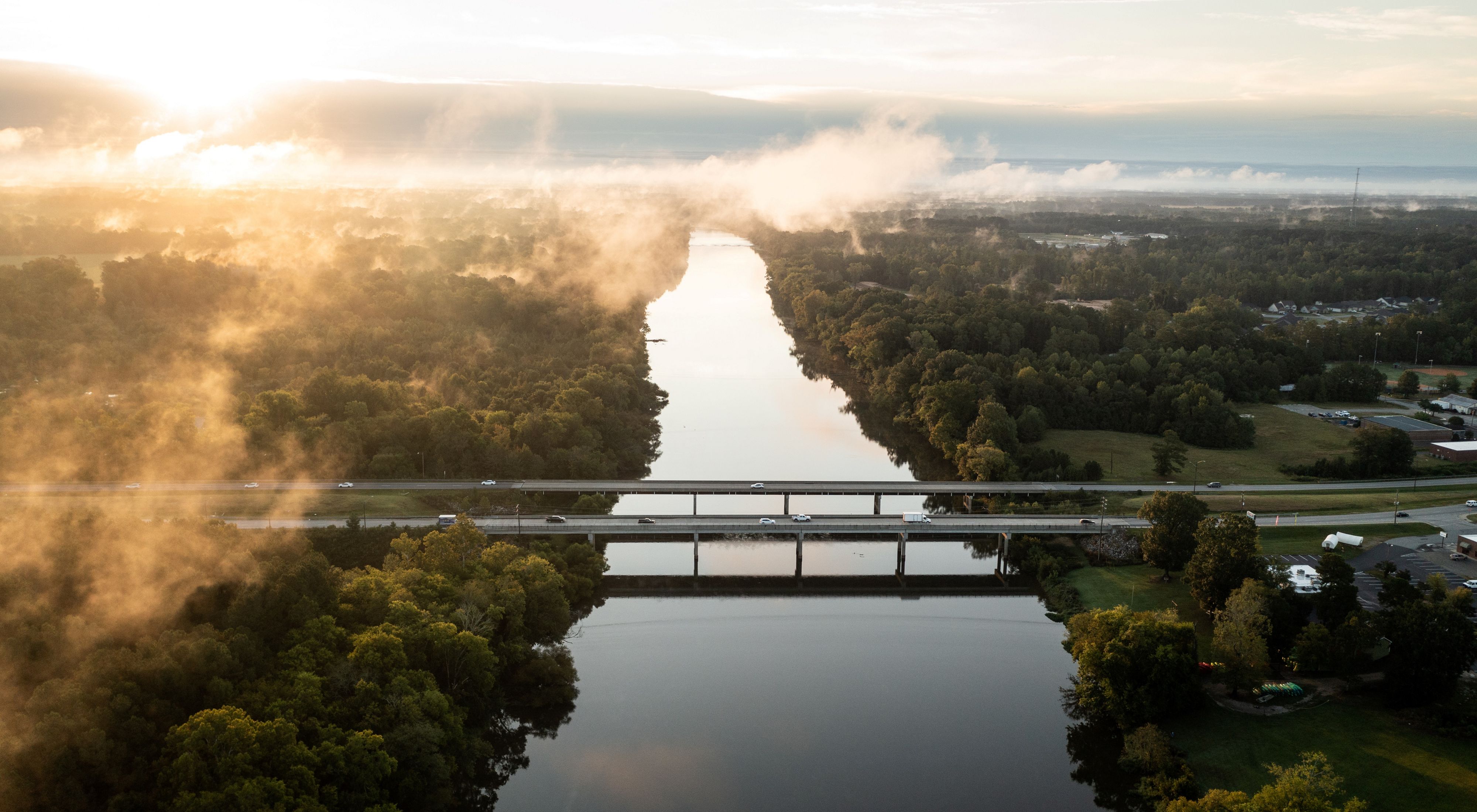 Sunrise over the Cape Fear River. Bridges carry cars across the river as white mist rises from the trees.