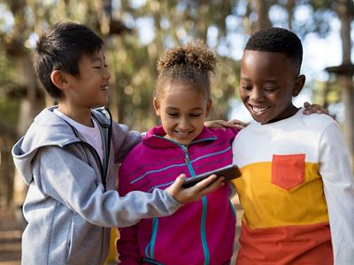 Three smiling children look at a mobile phone together while outdoors.