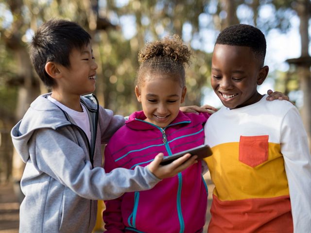 Three smiling children look at a mobile phone together while outdoors.
