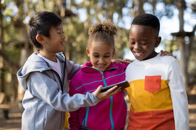 Three laughing children stand together looking down at a digital device that one of them is holding.