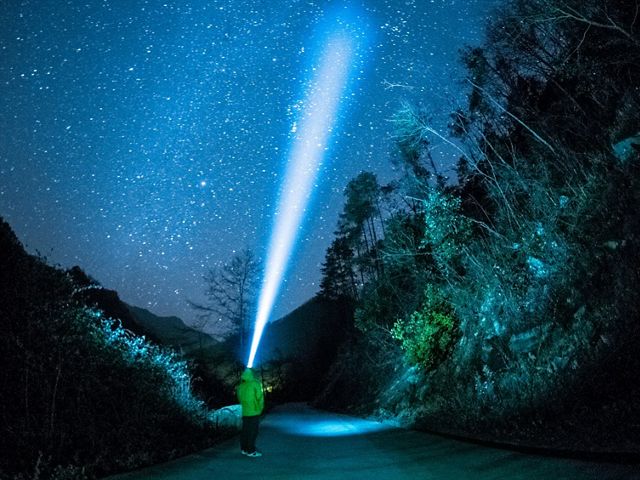 A person stands in a forest at night and shines a beam from their bright headlamp up into the night sky.