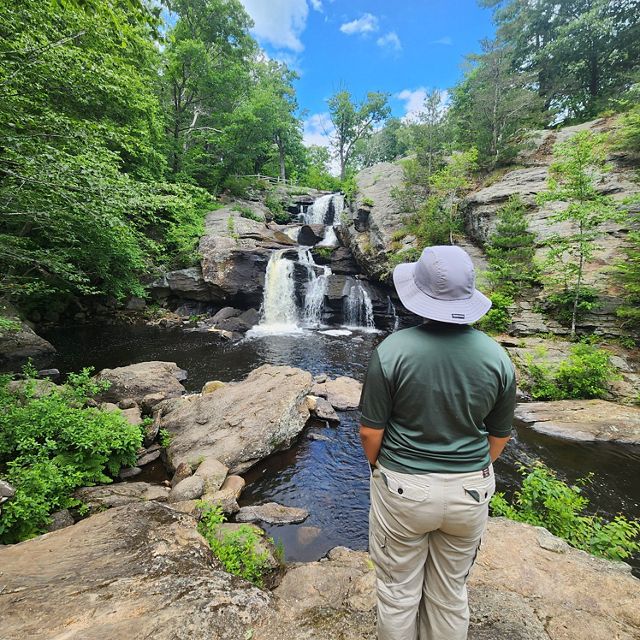 A person wearing a hat, and standing in front of a ravine, views a scenic waterfall surrounded by lush greenery and rocky terrain.