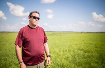 A man in sunglasses wearing a red shirt stands on the prairie. A blue, partly cloudy sky can be seen in the background.