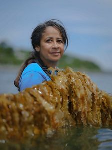 A woman stands holding a large strand of seaweed.