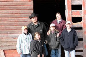A family of two parents and four kids stand in front of a barn.