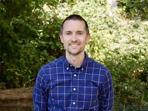 David Martin headshot. A smiling man wearing a blue plaid shirt poses outdoors in front of a tall green hedge.