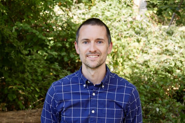 David Martin headshot. A smiling man wearing a blue plaid shirt poses outdoors in front of a tall green hedge.