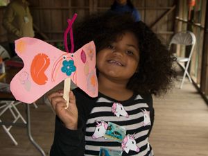 A little girl holds up a pink butterfly made of construction paper.