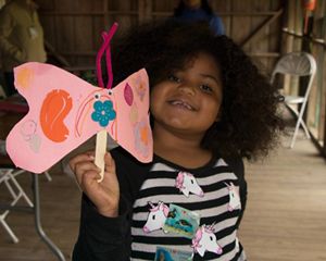 A little girl holds up a pink butterfly made of construction paper.