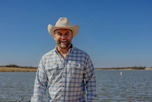 A man in a cowboy hat stands in front of blue coastal waters.