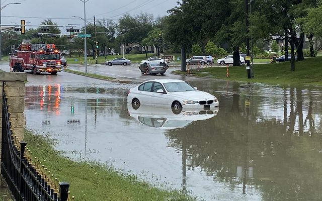 A car sits on a flooded street, stuck in high water, with a firetruck and other cars in the background.