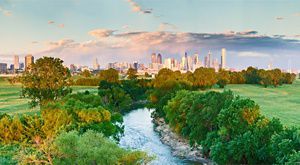 River and green floodplain in the foreground with the Dallas skyline glowing in evening light beneath dramatic clouds.