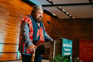 Dallas smith speaks into a podium while wearing traditional blue and red first nations garb and a a gray fedora
