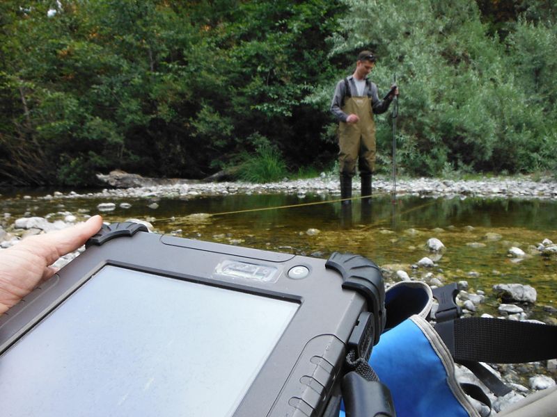 In the foreground, hands hold a digital tablet. in the background, a man wearing waders stands in ankle deep water collecting samples.