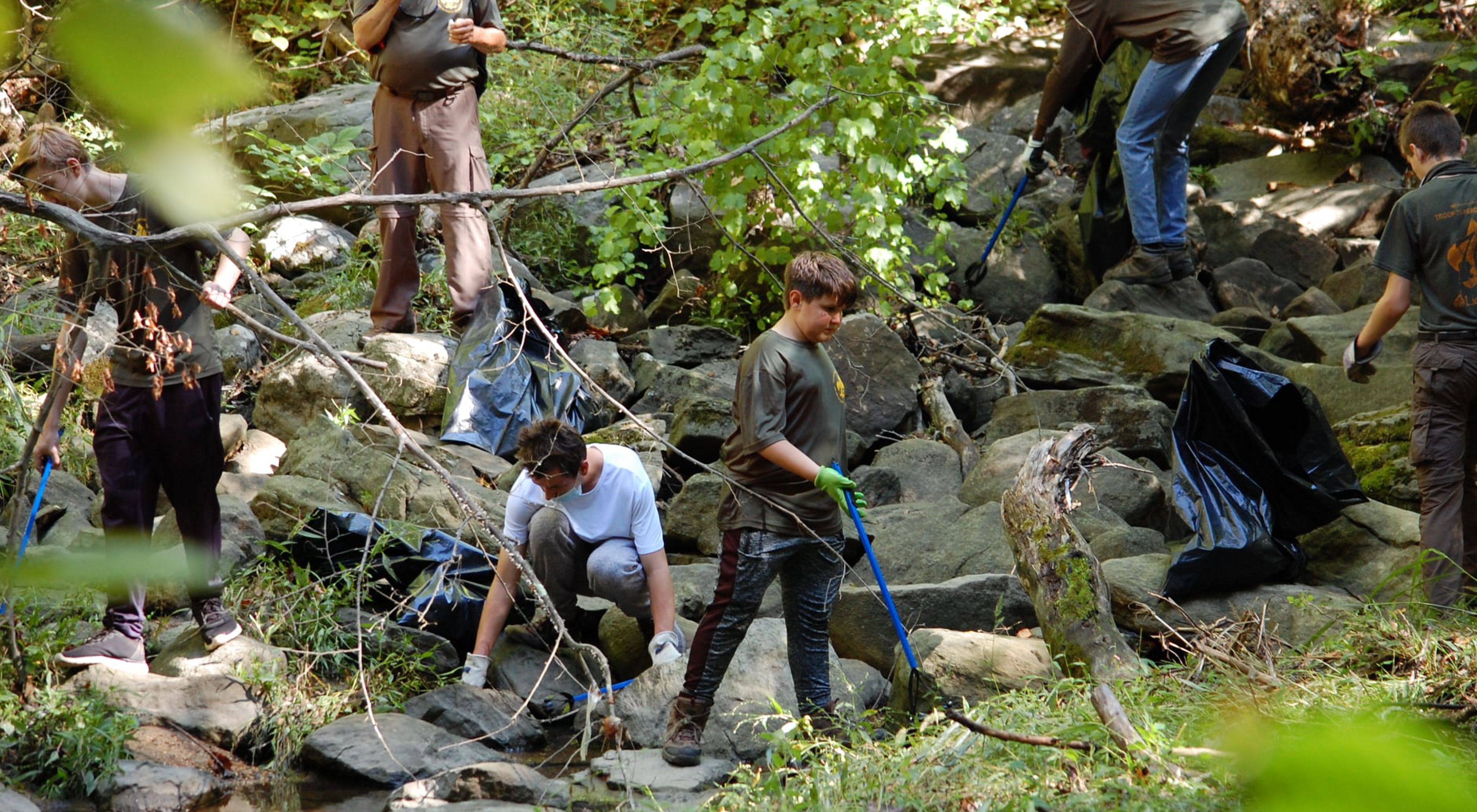 A group of adults and teens stand on rocks and boulders at the edge of a stream during a volunteer cleanup event.