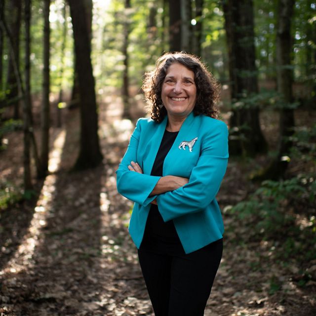 A woman with dark curly hair wearing a bright blue blazer stands in the forest with arms crossed.