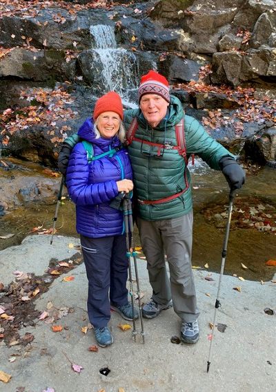 Deborah J. de la Reguera and her husband stand in front of a small waterfall and smile for the camera.