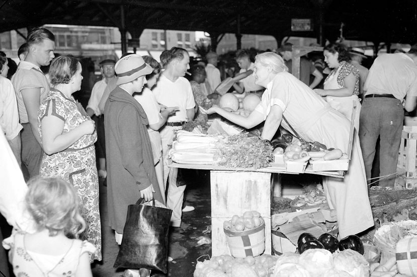 Black and white photo of a crowded market, with a vendor leaning over a table of produce for sale.