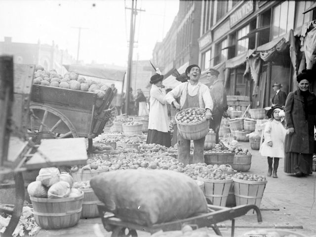Black and white photo of a man on a sidewalk holding a large basket of apples, surrounded by more baskets at his feet.