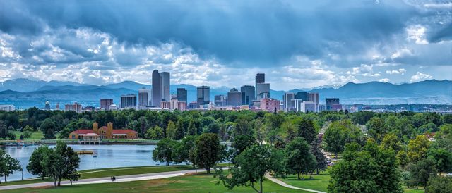 Landscape view of a green park filled with trees and a pond with the Denver skyline in the background.