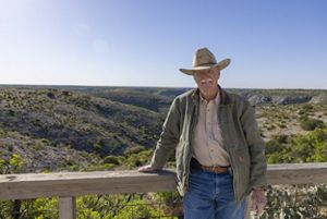 A person wearing a hat and jacket leans on a wooden railing at an overlook, with expansive rocky hills and green valleys stretching into the distance behind them.