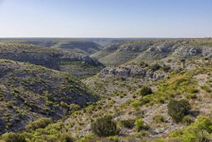 A panoramic view features a rocky canyon carved into rolling hills, dotted with shrubs and grasses, stretching toward the horizon under a bright blue sky.