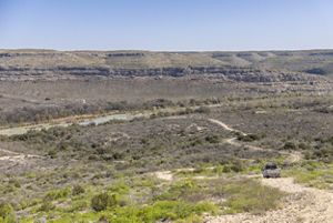 A wide view features a rugged landscape with low hills, sparse vegetation and a river visible in the distance, while a pickup truck drives along a dirt road in the foreground.