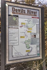 A large informational sign titled “Devils River Mile 12 Paddler Camp” shows a detailed map of a natural area with marked hazards, access points and camp rules, mounted outdoors among trees and dry grass.
