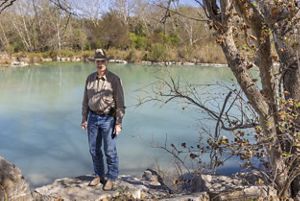A person wearing a cowboy hat, button‑down shirt, jeans and boots stands on a rock ledge beside still, turquoise river water, with trees and brush lining the opposite bank.