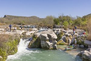 A group of people stand and move carefully across large limestone rocks at a shallow rapid, while canoes are pulled through clear, fast‑moving water surrounded by trees and brush.