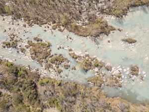 An overhead view features pale green river water splitting into multiple shallow channels around rocky outcrops and small islands of brush and trees.
