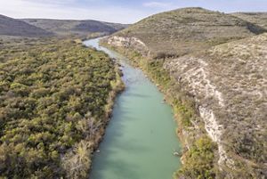 An aerial view features a river winding through a narrow limestone canyon, with pale turquoise water bordered by dense green shrubs on one side and rocky, tan hills on the other.
