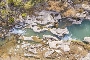 Aerial view of a turquoise river flowing through smooth, pale limestone formations dotted with shallow pools, surrounded by dry grasses and scattered green shrubs.